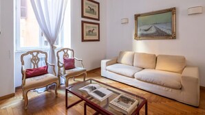 A living room with a beige sofa, two wooden chairs with cushions, a glass coffee table with books, a painting on the wall, and a window with sheer curtains. - Virgilio by Interhome (Roma: Piazza Navona - Campo dei Fiori)