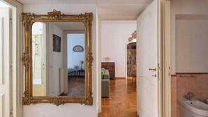 A hallway with a large ornate mirror, wooden flooring, a door leading to a bathroom with a bathtub, and a glimpse of a bedroom with a bed and a lamp. - Virgilio by Interhome (Roma: Piazza Navona - Campo dei Fiori)