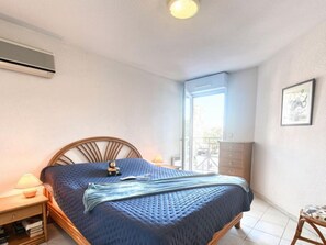 A bedroom with a bed, bedside table, lamp, air conditioning unit, and a view through a window.