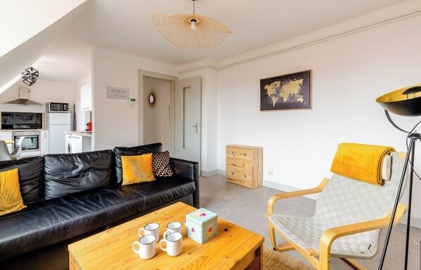 A modern living room with a black leather sofa, a wooden coffee table with mugs, a yellow armchair, and a kitchen in the background. - 2-room apartment. Right in the historic city center! (Sélestat)