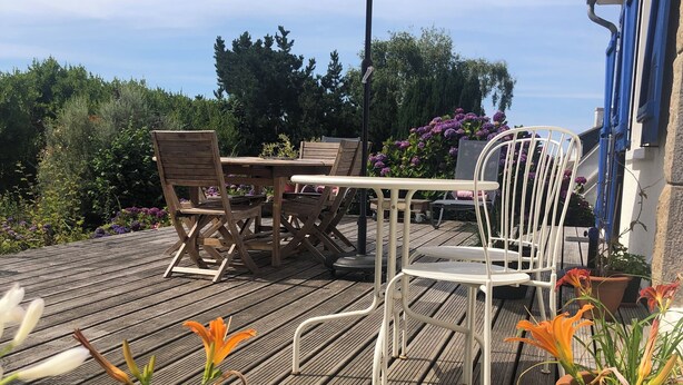 Patio esterno con pavimento in legno, un tavolo, sedie, un ombrellone grande e piante in vaso.
