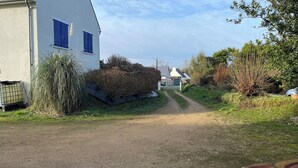 Un sentiero stretto in ghiaia conduce a un edificio bianco con persiane blu, immerso nel verde e con una piccola costruzione bianca sullo sfondo.