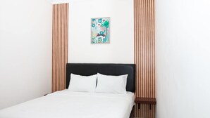 Un lit bien fait, avec du linge de lit blanc, une tête de lit sombre et une petite table de chevet surmontée d’une lampe.