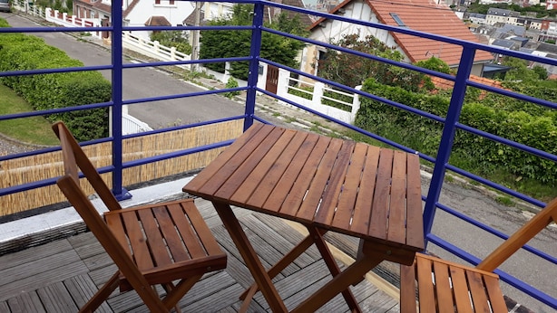A balcony with a wooden table and chairs, overlooking a residential area with houses and a view of the sea.