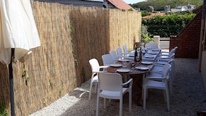 An outdoor patio with a table set for dining, surrounded by white chairs, a bamboo fence, and a view of a hill with buildings.