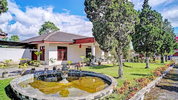 A traditional house with a tiled roof, a small garden with a circular pond, and a row of trees. - Capital O 92214 Beristera Hotel (Kota Sidikalang)