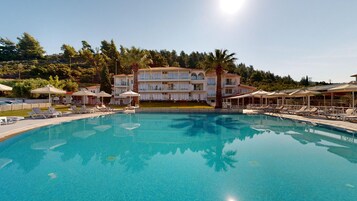 A swimming pool with lounge chairs and umbrellas, a multi-story building with balconies, and a forested hillside in the background.