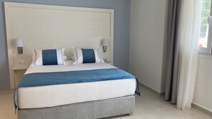 Una cama bien hecha con base gris y ropa de cama azul, flanqueada por dos lámparas de noche, en una habitación con un cabecero blanco sencillo y una ventana con cortinas.