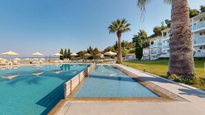 A swimming pool with lounge chairs and umbrellas, surrounded by palm trees and a building with balconies. - Acrotel Lilyann Boutique Hotel (Sithonia)