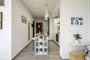 A modern hotel corridor with a white reception desk, a seating area with chairs, a white shelving unit with plants, and a hanging pendant light.