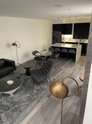 Un salon moderne avec un canapé, une table basse ronde et un fauteuil noir. On y trouve également une table à manger avec des chaises, un coin cuisine avec un réfrigérateur et un évier, ainsi qu’une grande fenêtre.