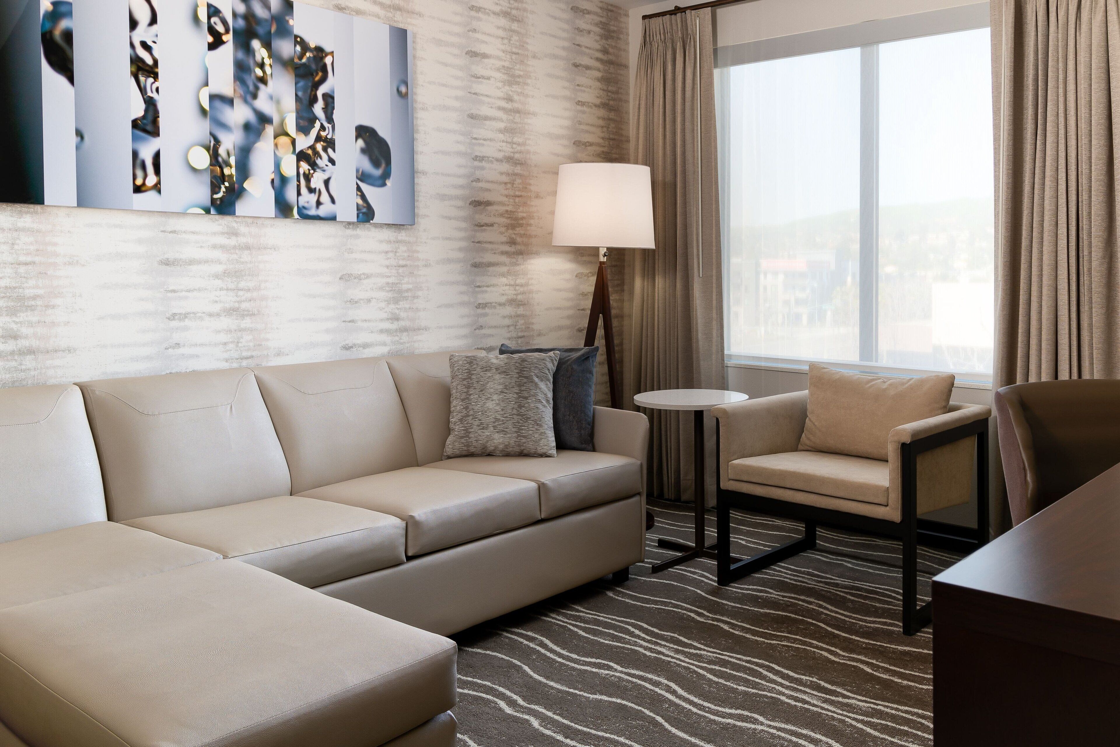 Una habitación de hotel moderna con un sofá color beige, una mesa redonda pequeña, una silla y un ventanal con cortinas.