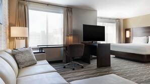 A hotel room with a sofa, a desk with a flat-screen TV, a bed, and large windows with curtains. - Residence Inn by Marriott Anaheim Brea (Brea)
