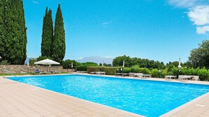 Una piscina rectangular con agua azul cristalina, rodeada por una terraza con baldosas. Hay varias sillas de descanso y sombrillas a lo largo de la piscina. Delgados árboles altos bordean el fondo, y un muro de piedra separa la zona de la piscina de la vegetación.