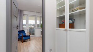 Una habitación con piso de madera, una silla azul, un armario blanco con puertas de vidrio y una ventana con vistas a edificios.