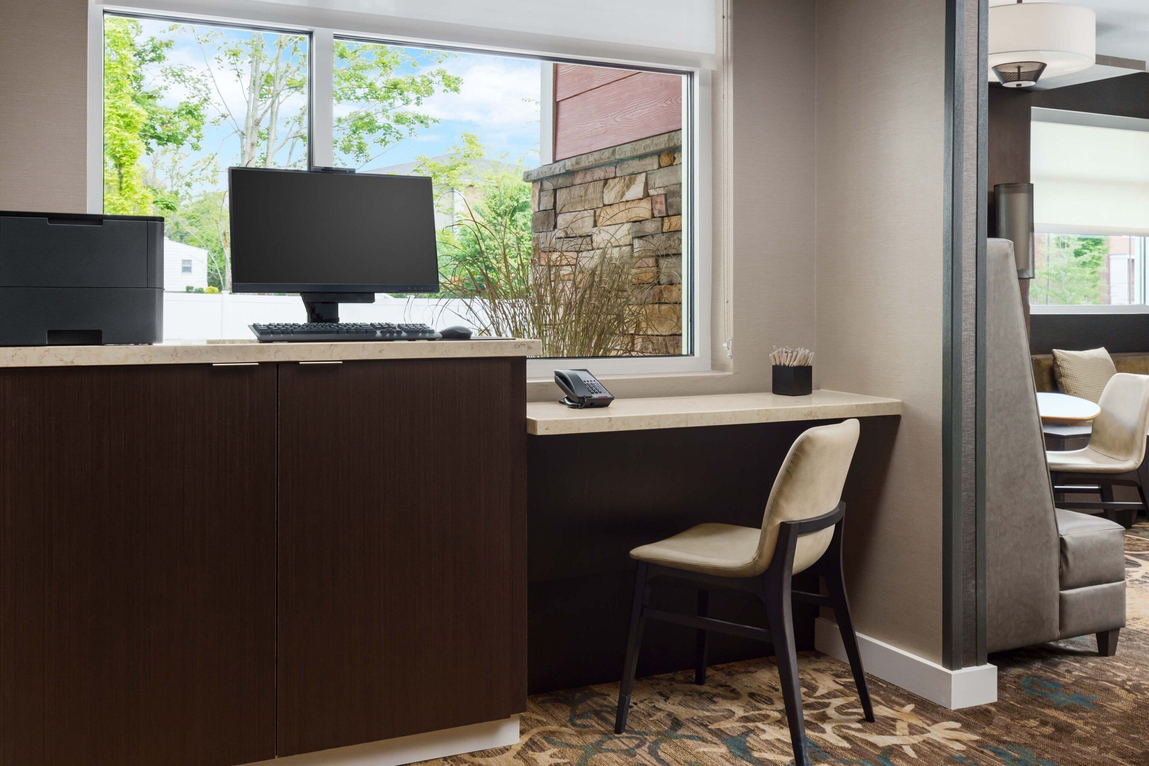 A hotel room with a desk, computer, and chair, a window with a view of trees, and a patterned carpet.