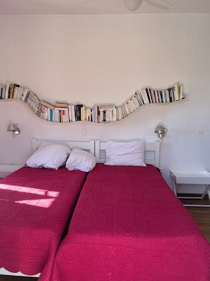 Un dormitorio con dos camas, una estantería, dos lámparas de noche y una colcha roja.