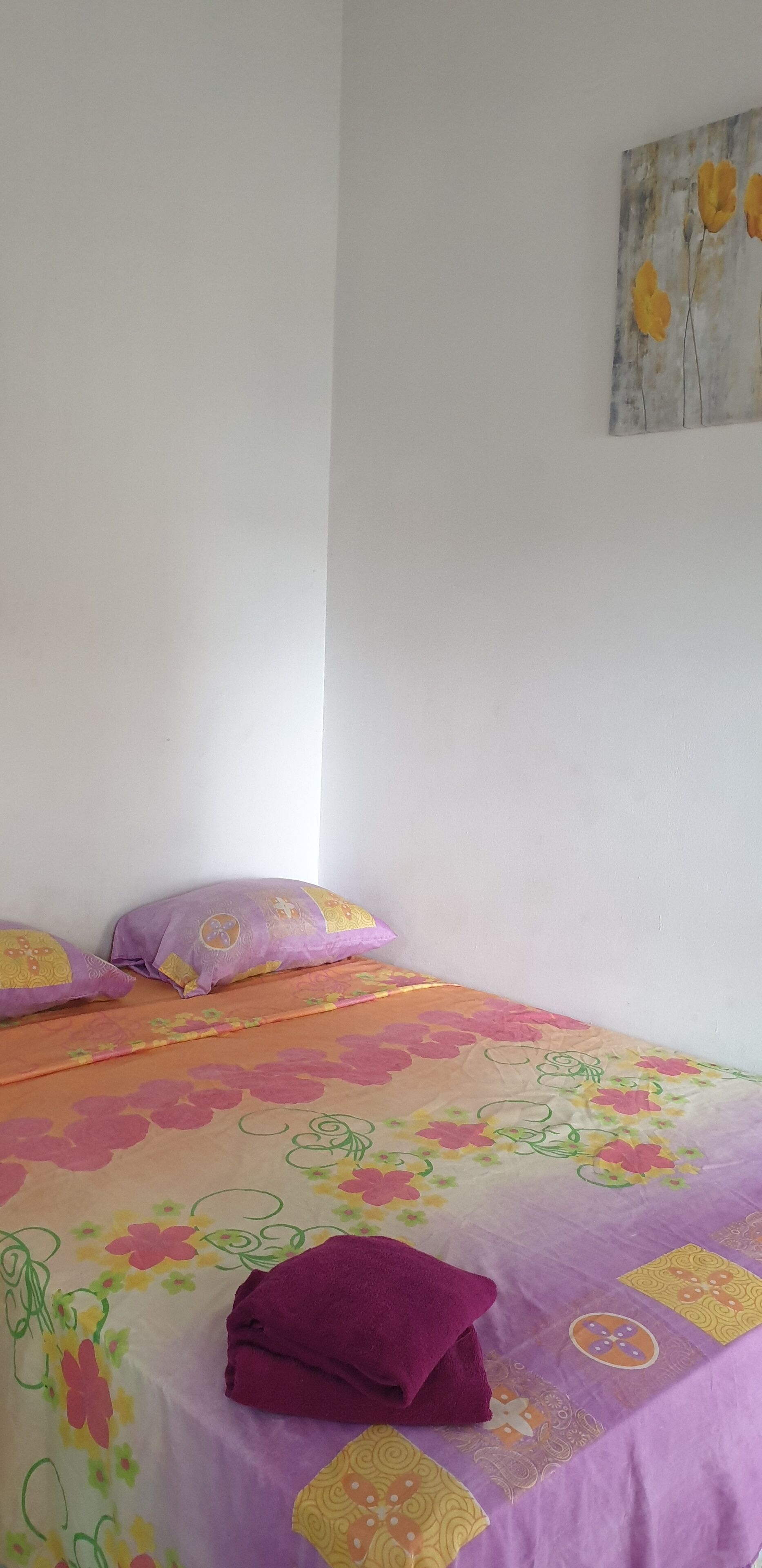 A bed with a floral bedspread and two purple pillows, a folded purple towel, and a painting of flowers on the wall.