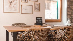 Un comedor con mesa de madera y sillas tejidas, obra de arte enmarcada en la pared y ventana con vista a un edificio de piedra.