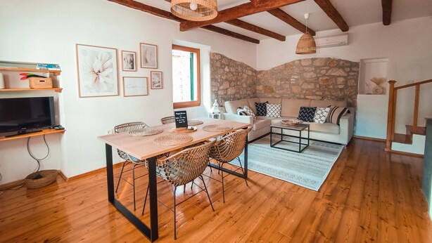 Una sala de estar acogedora con pared de piedra, vigas de madera, un comedor con sillas de mimbre y un sofá con cojines estampados.