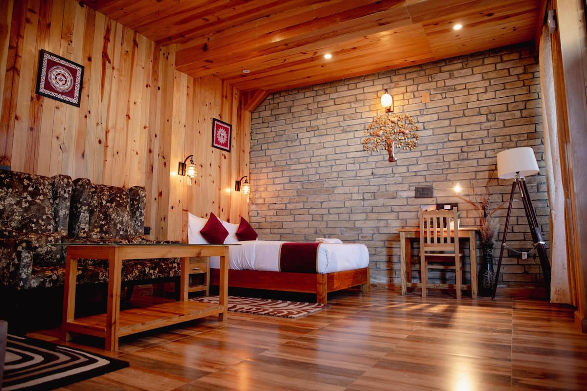 A cozy room with a wooden ceiling, a brick wall, a bed with red bedding, a wooden sofa, a small table, and a lamp.