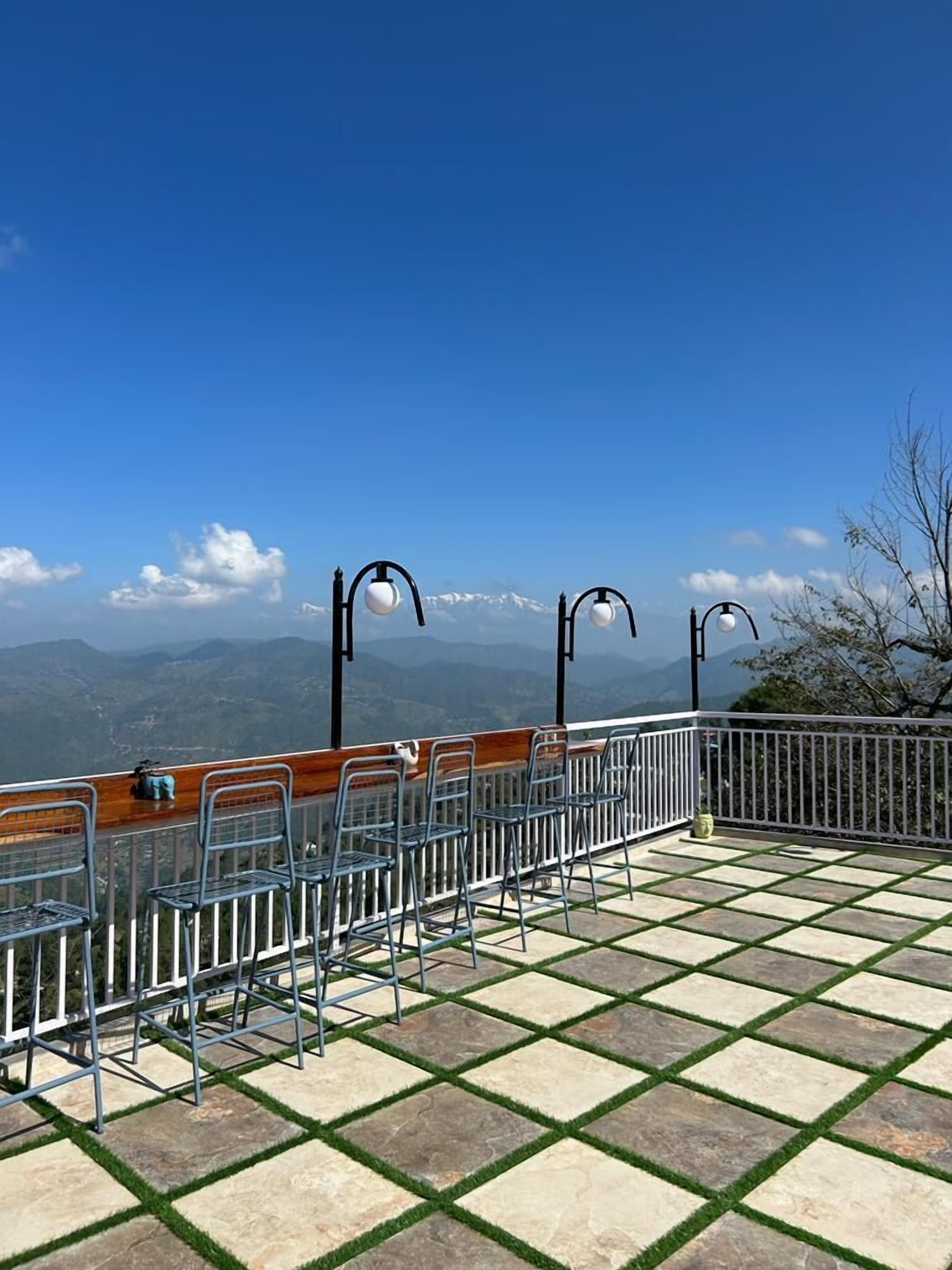 Een terras met uitzicht op bergen, metalen stoelen, een houten tafel en een hekwerk.