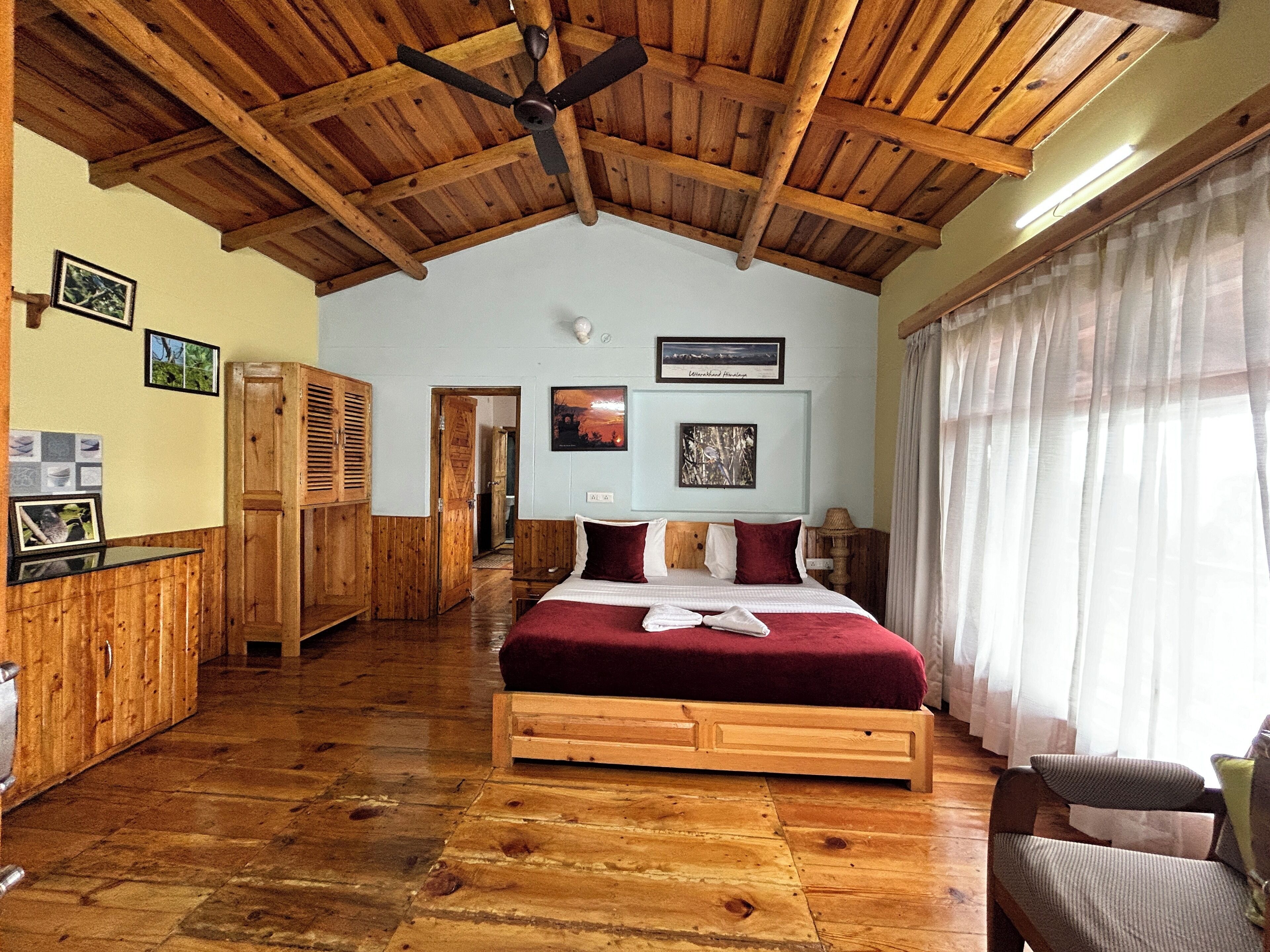 Una habitación con piso de madera, una cama, un ventilador de techo y vistas al exterior a través de una ventana con cortinas translúcidas.