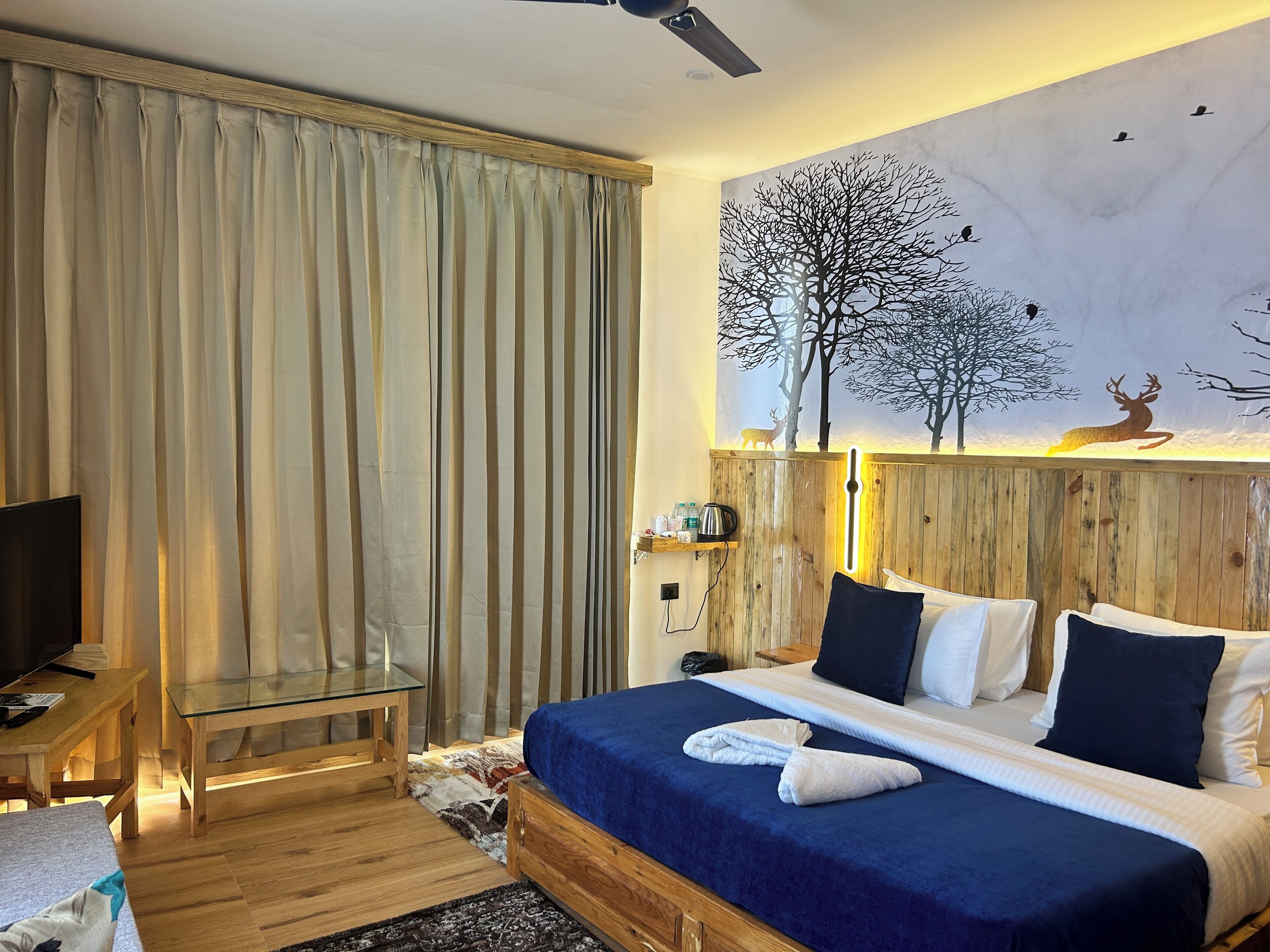 A hotel room with a large bed, a television, a wooden desk, and a painting of a tree and deer on the wall.