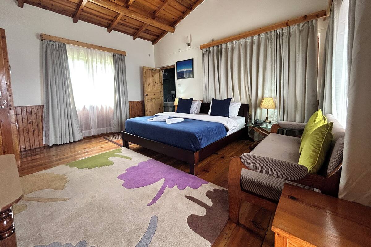 A bedroom with a wooden ceiling, a bed with blue bedding, a nightstand with a lamp, a chair, and a window with curtains.
