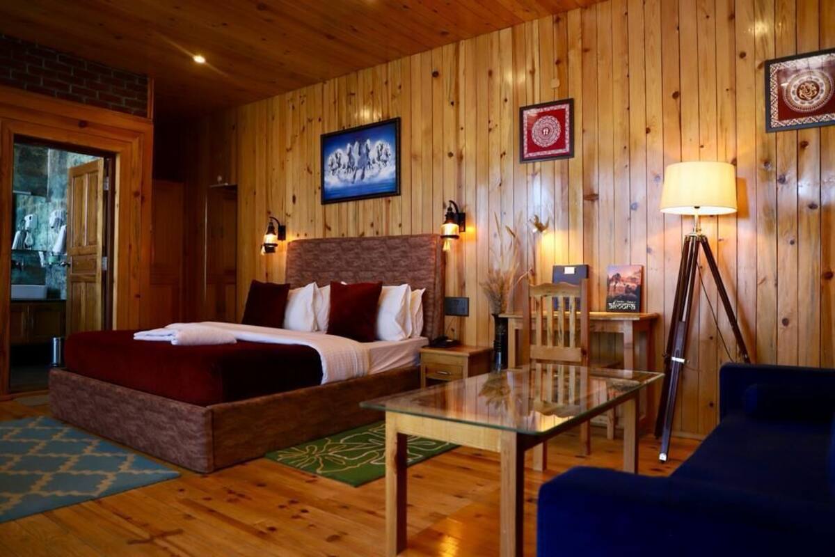 A wooden-paneled room with a bed, a glass table, a blue sofa, and a small table with a lamp.