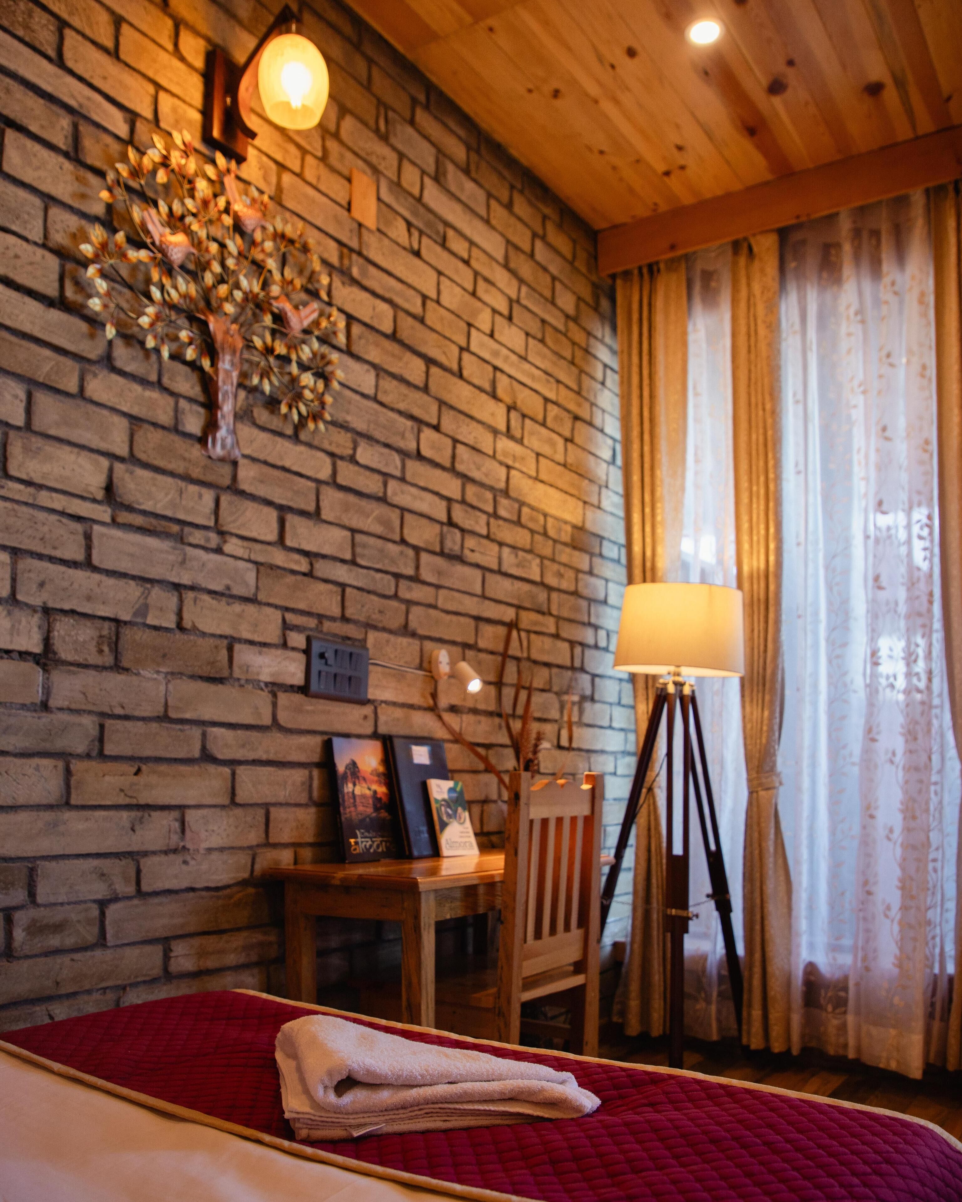 A room with a brick wall, a wooden ceiling, a bed with a red quilt, a lamp, a chair, and a small table with a picture frame and a book.