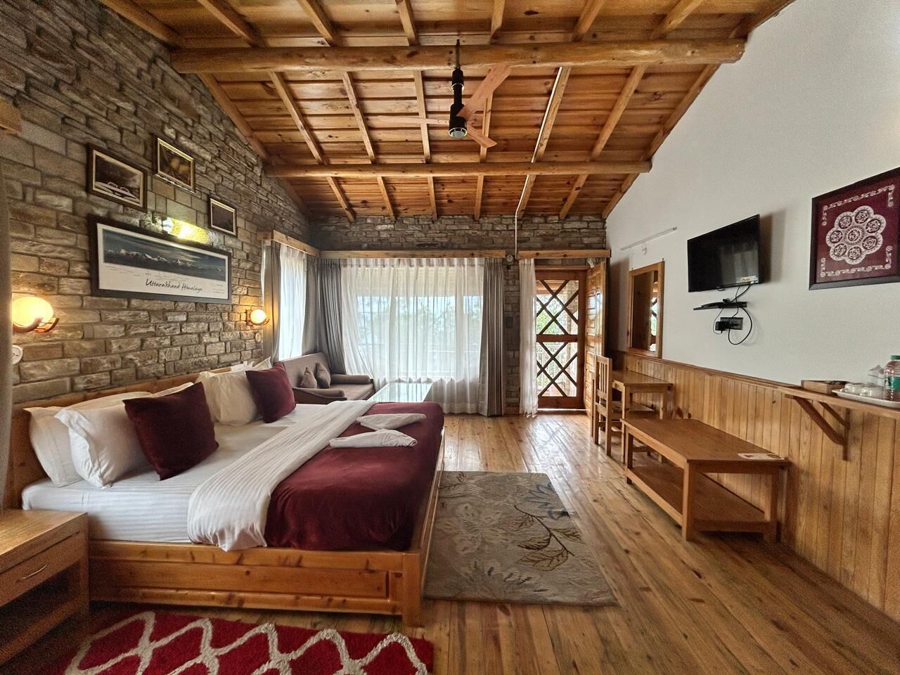 A cozy room with a stone wall, wooden ceiling, a bed with a red bedspread, a wooden bench, a TV, and a window with curtains.