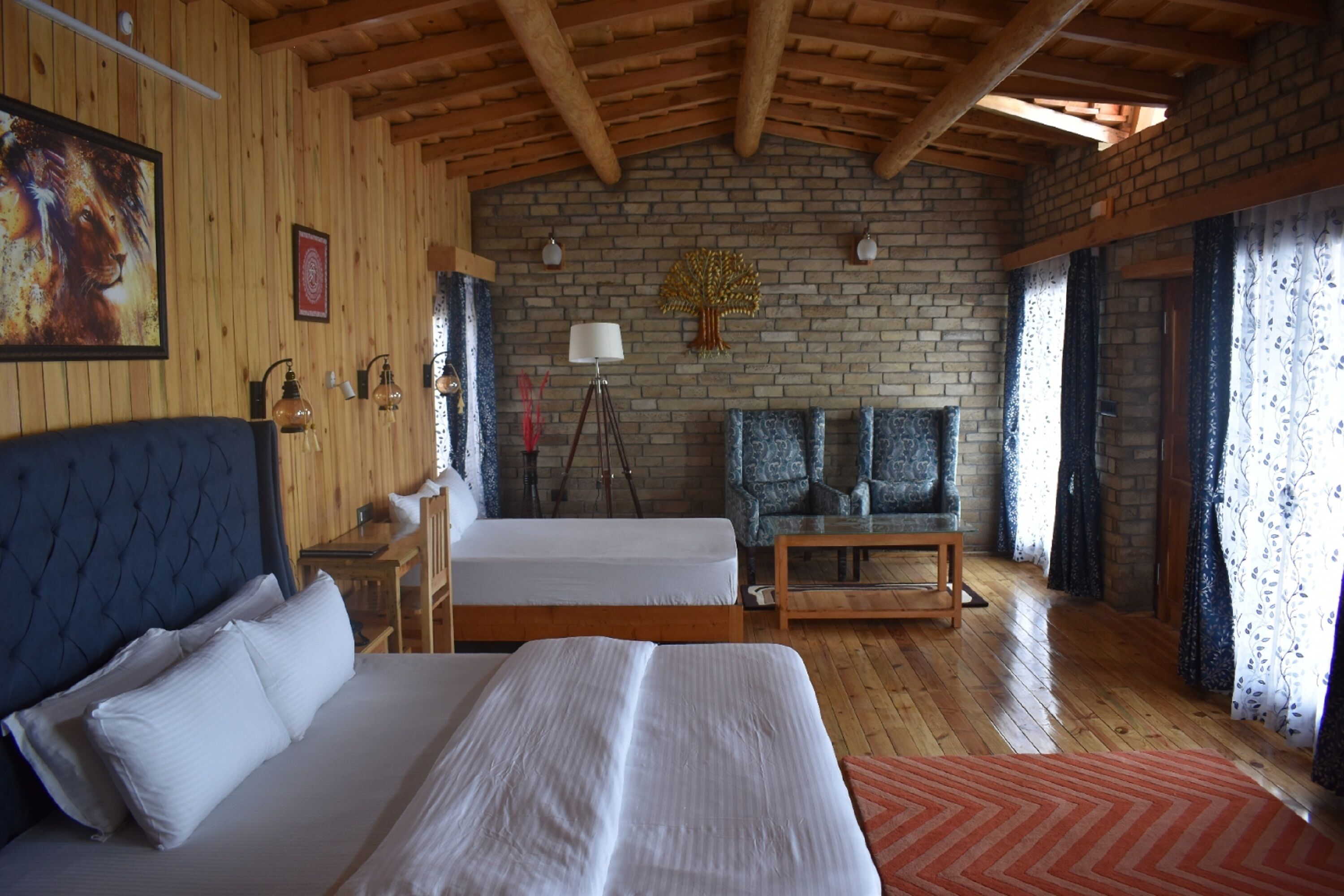 A room with two beds, a wooden ceiling, a brick wall, and a large window with curtains.