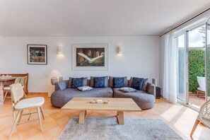 Un salon moderne comprenant un canapé d’angle, une table basse en bois, un coin repas avec une table et des chaises, et une grande fenêtre donnant sur un paysage verdoyant.