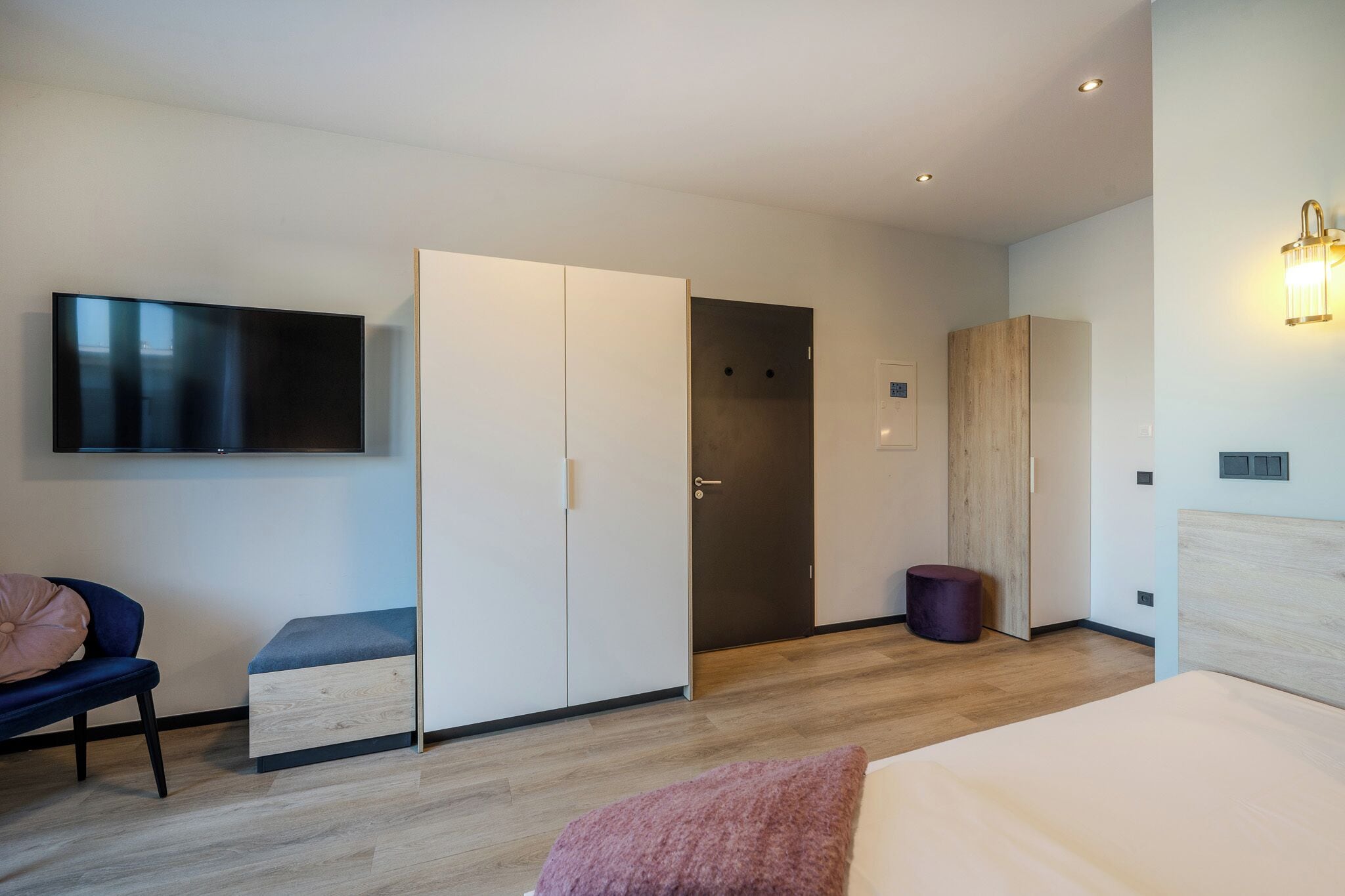 Ein modernes Hotelzimmer mit Flachbildfernseher, Kleiderschrank, Bett und einer kleinen Sitzecke.