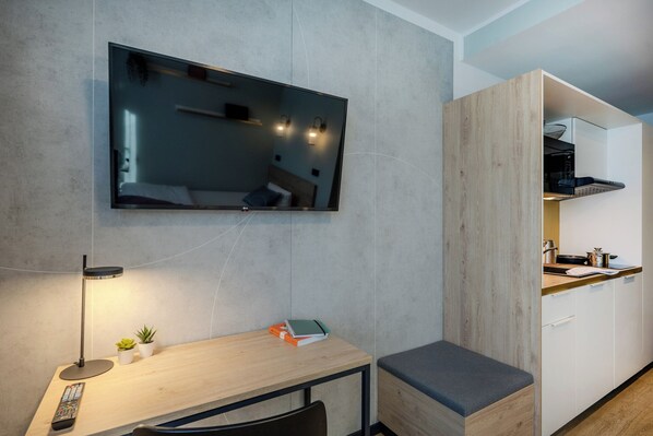 A modern room with a flat-screen TV mounted on the wall, a wooden desk with a lamp, a small plant, and a remote control. - The Base Berlin ONE (Berlin)