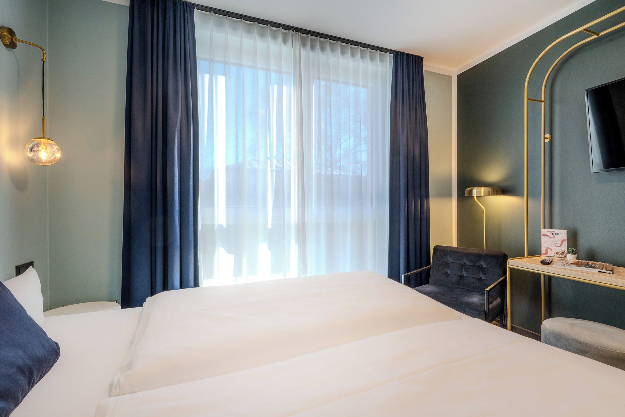Habitación de hotel con una cama grande, un escritorio con silla, un televisor montado en la pared y una ventana con cortinas.