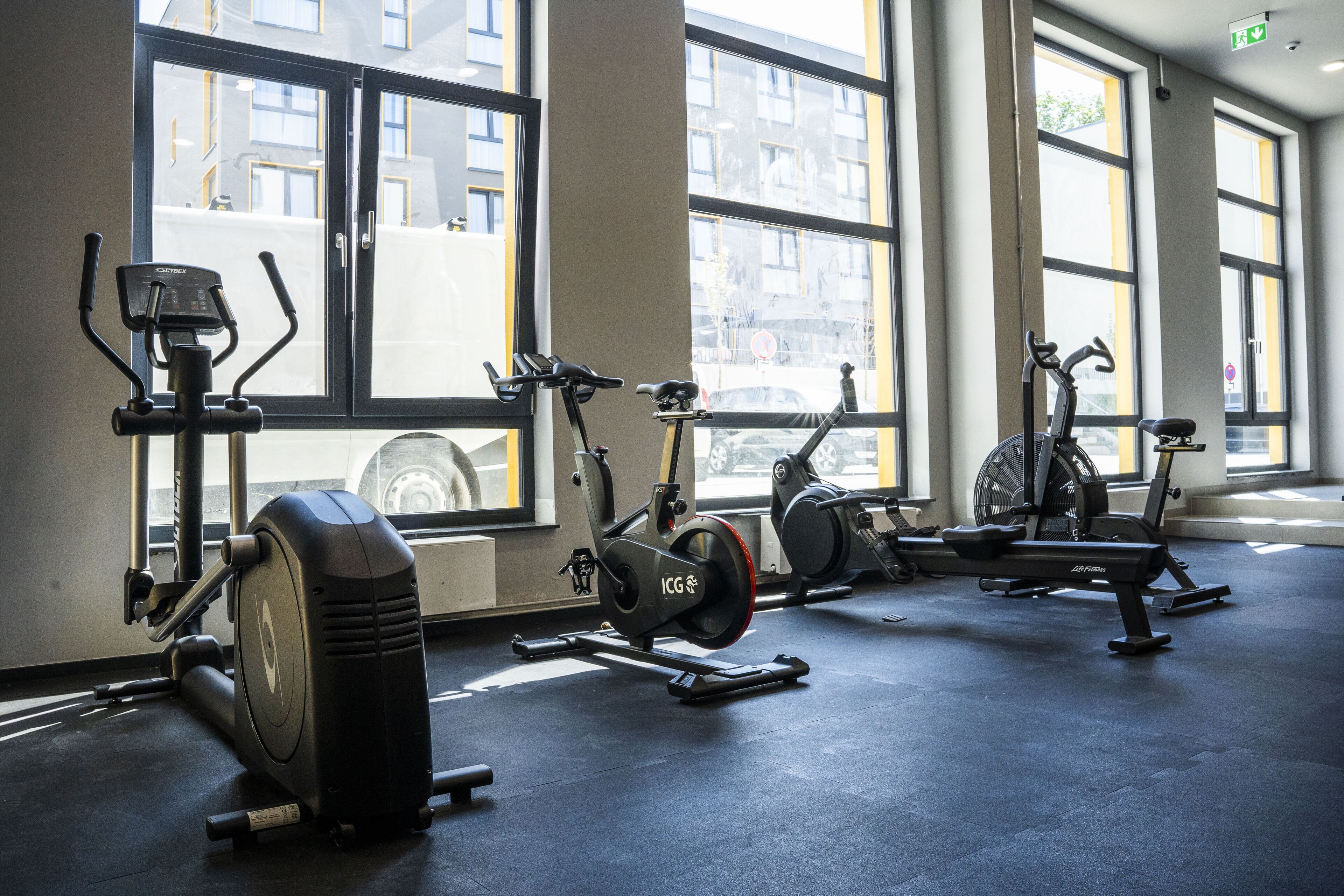 Ein Fitnessraum mit Ergometer und Crosstrainern, großen Fenstern und einem sauberen Boden.