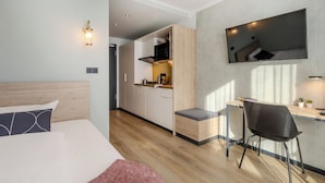 A modern hotel room with a bed, a desk, a kitchenette, and a TV mounted on the wall. - The Base Berlin ONE (Berlin)