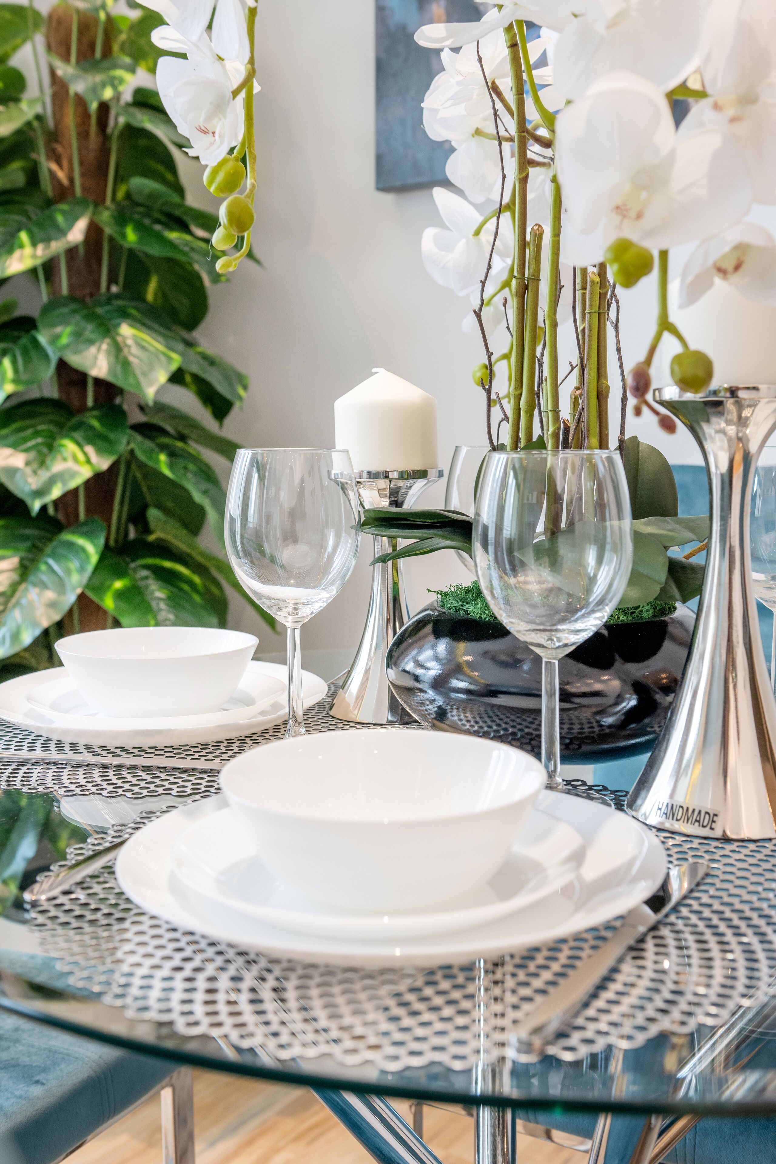 A dining table set with white plates, silverware, and clear glassware, surrounded by lush green plants and white orchids.