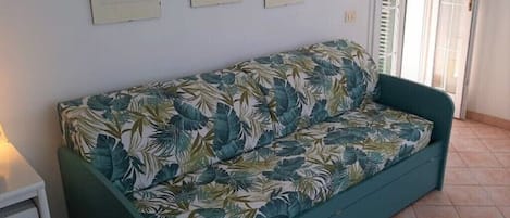 A teal sofa with a tropical leaf pattern, a small white bedside table, and a window with white shutters.