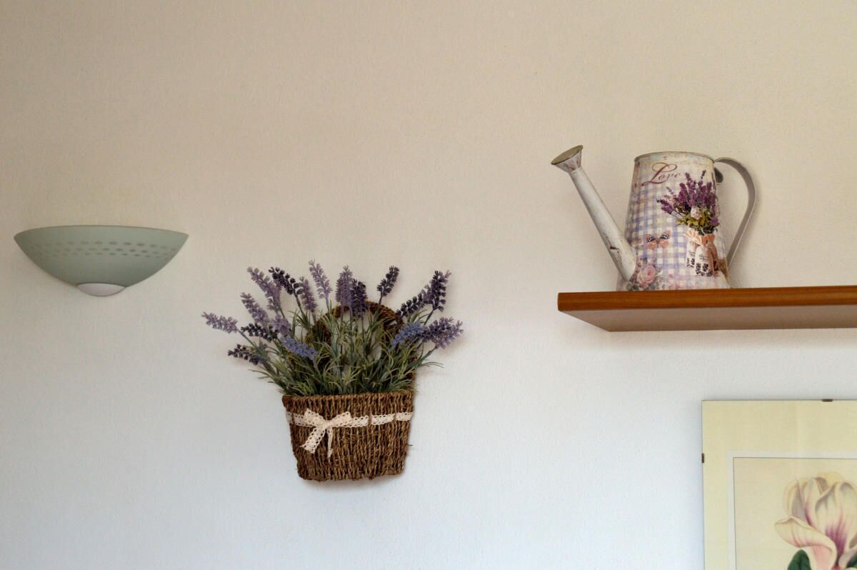 Pared con una cesta tejida que contiene lavanda, un aplique de pared y un estante con una regadera decorativa.