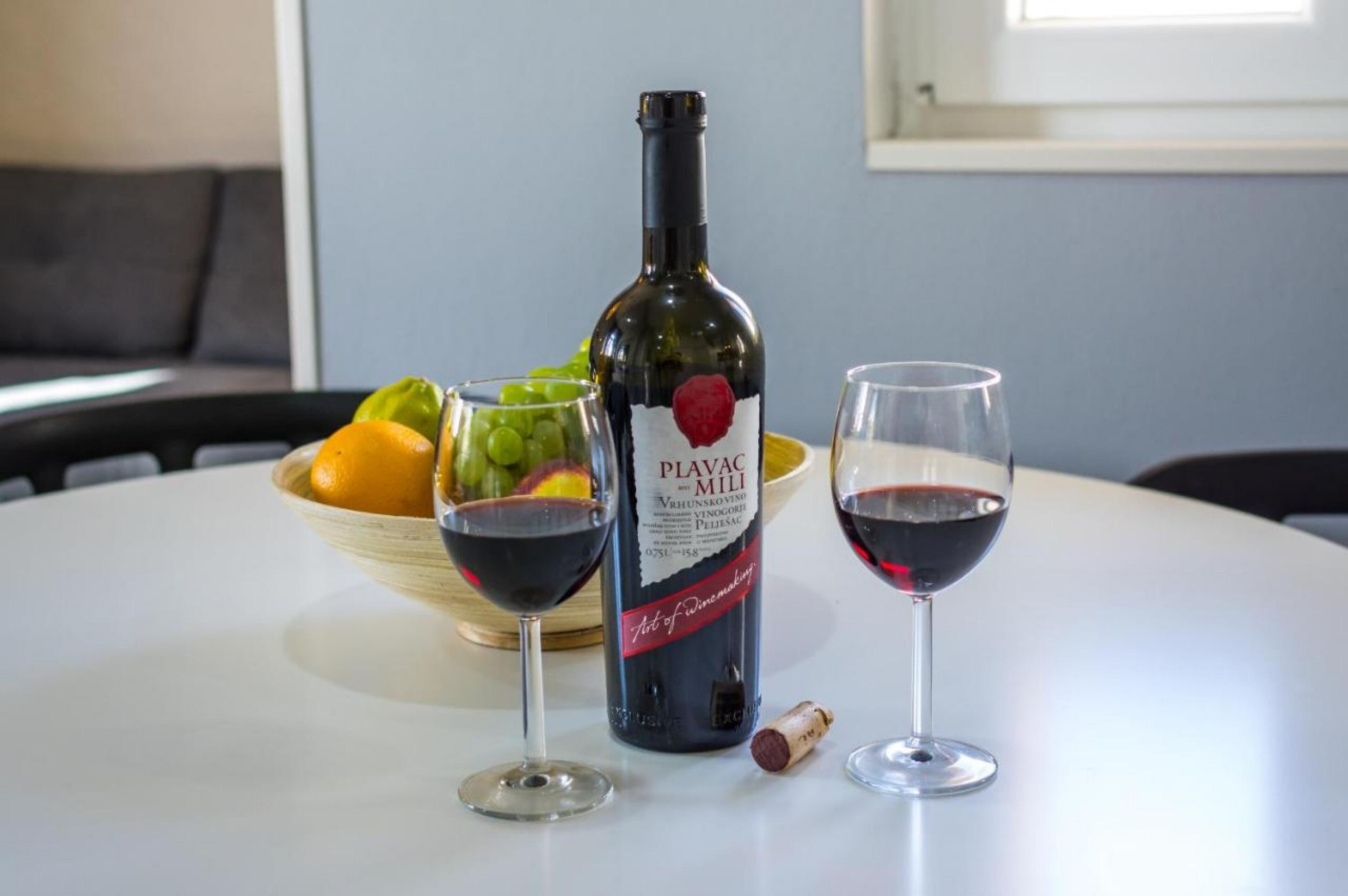 A bottle of Plavac Mili wine, two glasses of red wine, a bowl of fruit, and a cork on a white table.