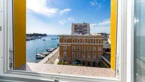 Blick aus einem Fenster auf einen Yachthafen mit Booten, einem gelben Gebäude und einem mehrstöckigen Bau im Hintergrund.