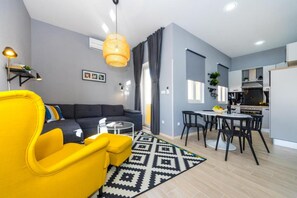 En moderne lejlighed med en stue, der indeholder en gul lænestol, en mørk sofa og et sofabord i glas. Køkkenet har en hvid bordplade, sorte stole og et spisebord.