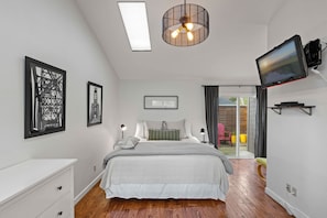 Une chambre à coucher comprenant un lit, des tables de chevet, un luminaire au plafond, un téléviseur fixé au mur et une porte-fenêtre coulissante donnant sur un espace extérieur.