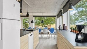 A modern kitchen with a built-in refrigerator, wooden cabinets, a sink, and a coffee machine. There are blue chairs around a table, and the room has large windows with a view of trees.