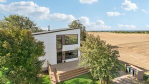 A modern two-story house with large windows, a wooden deck, and a view of a field.