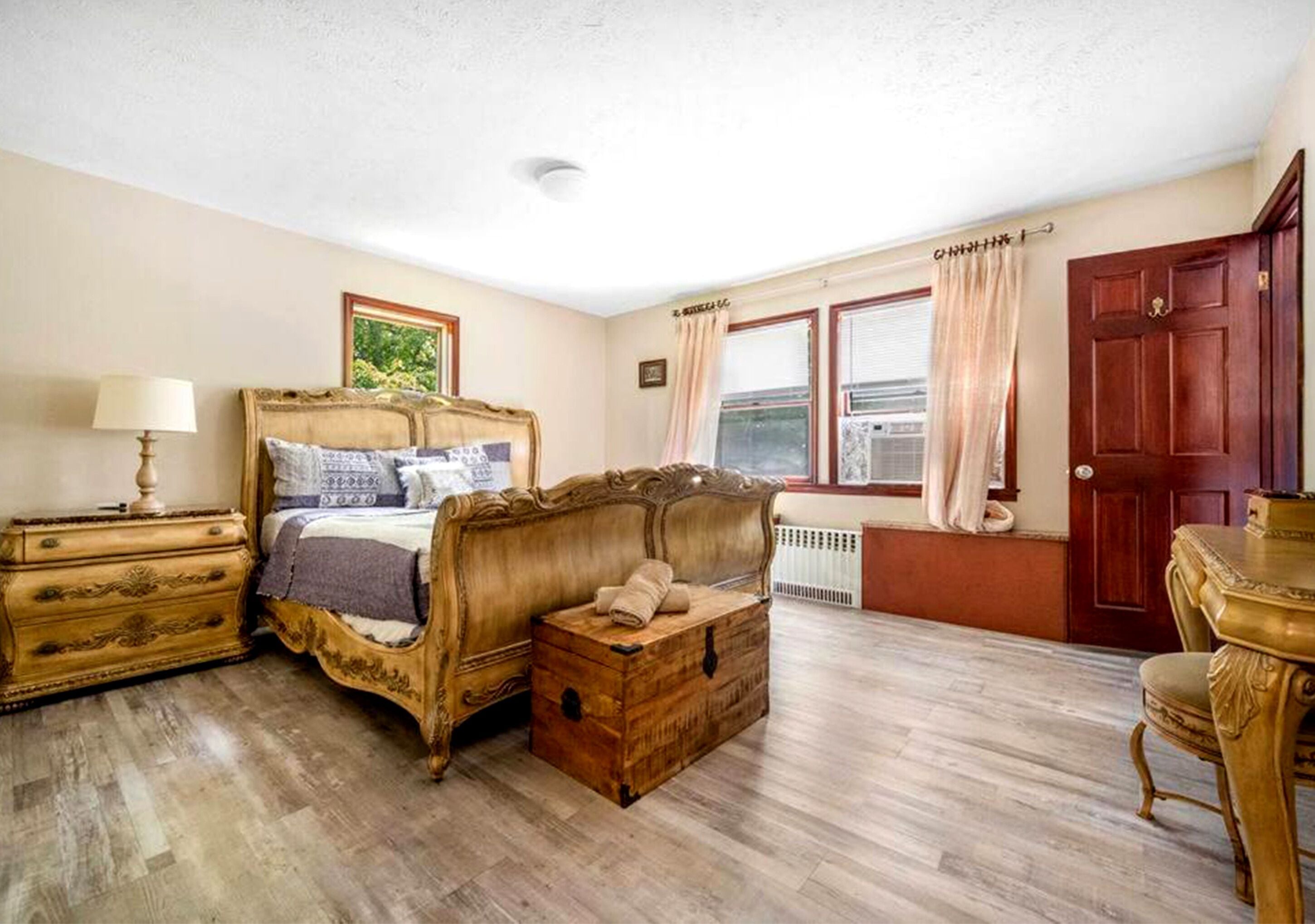 A bedroom with a wooden bed, a chest of drawers, a bedside table, a window with curtains, and a door.