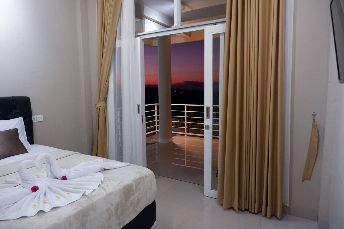 A hotel room with a bed, a balcony with curtains, and a view of the sunset.
